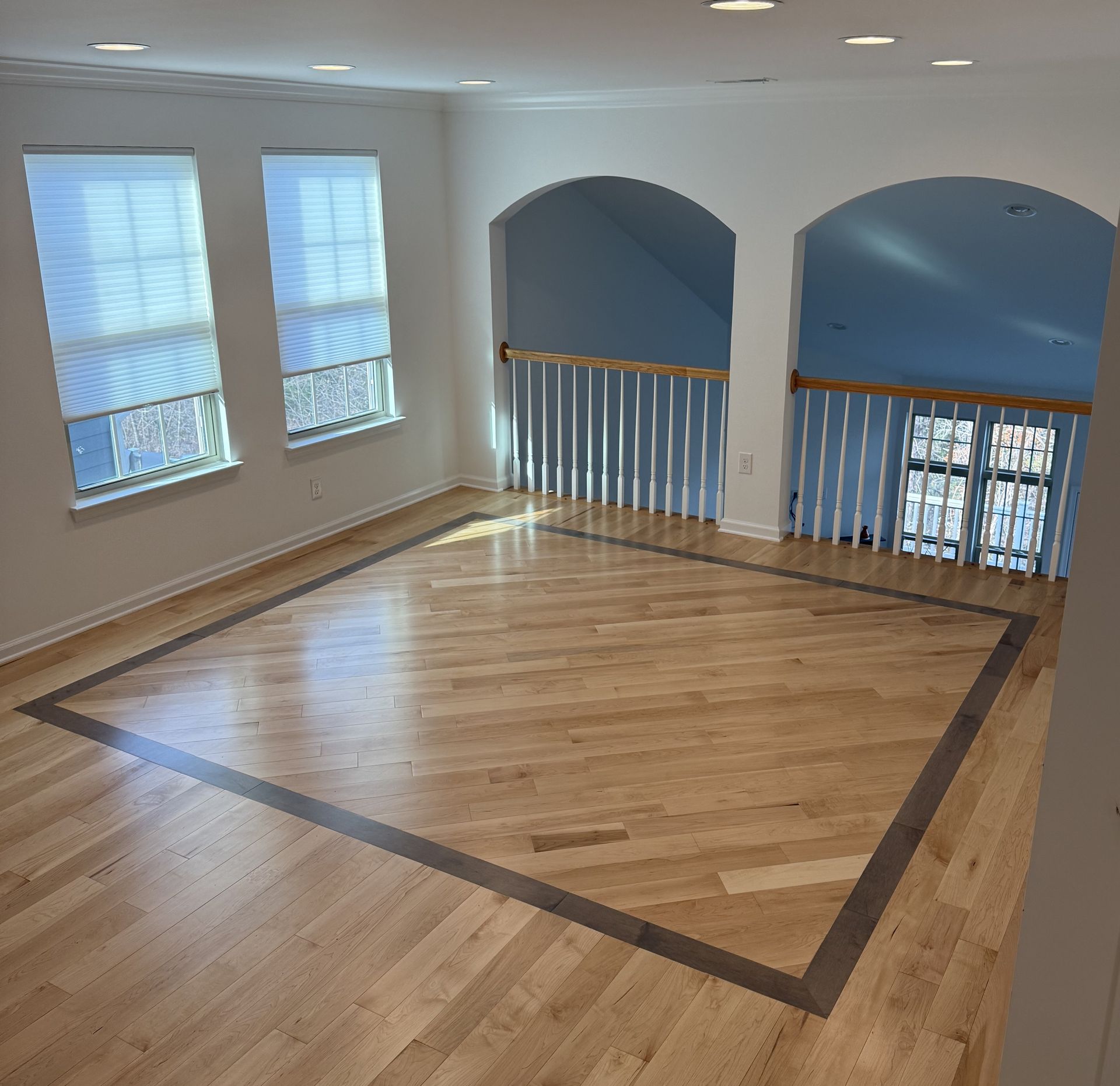 Empty hardwood loft room with windows, arched openings, and a dark border inlaid in the floor