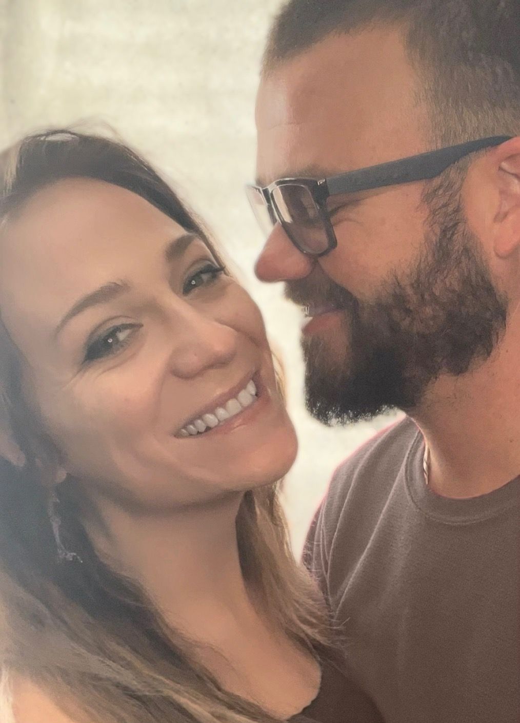 Two people smiling closely at each other indoors, with one wearing glasses and a beard.