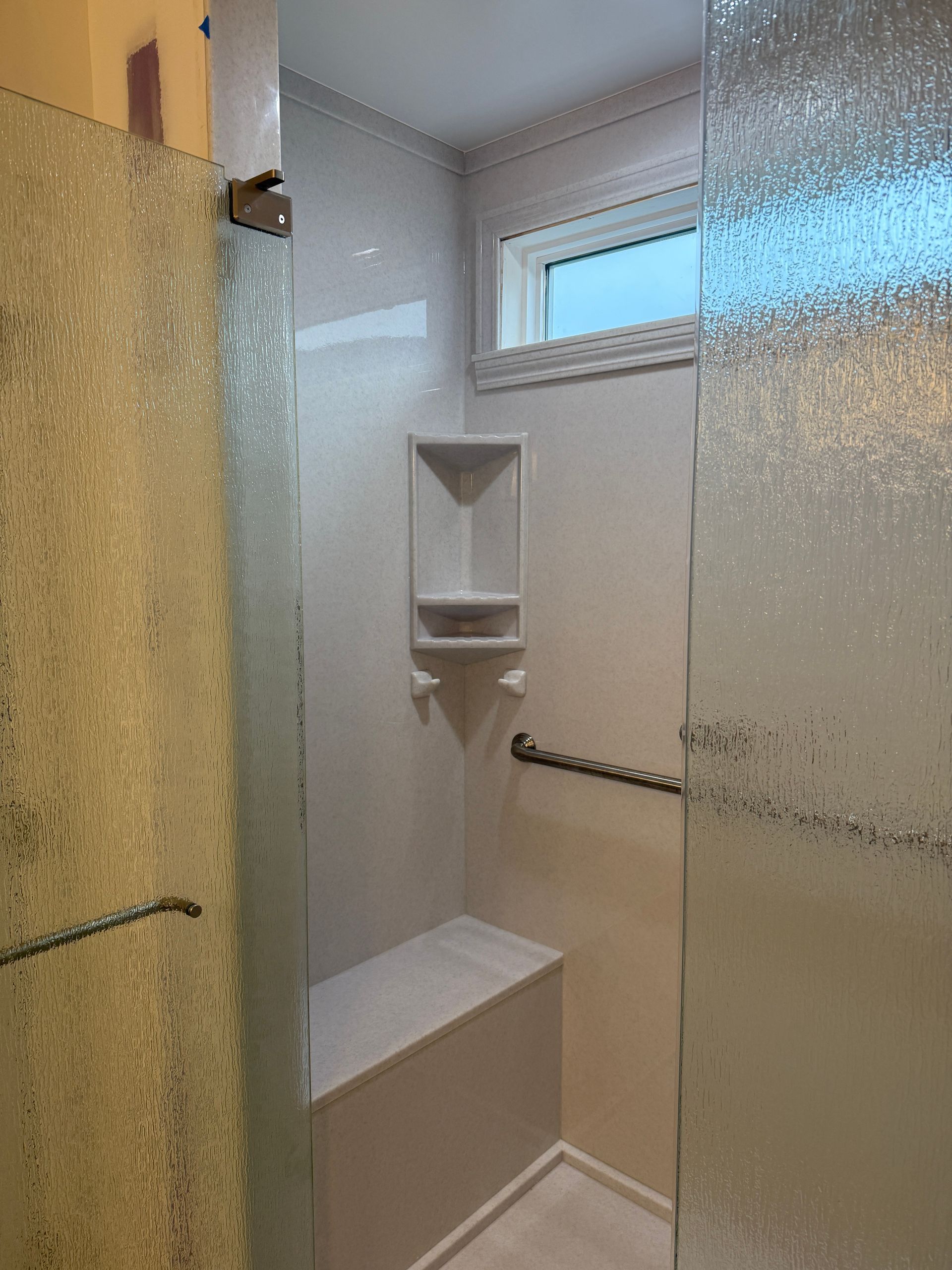 Small shower stall with frosted glass doors, built-in shelf, grab bar, bench, and a high window.