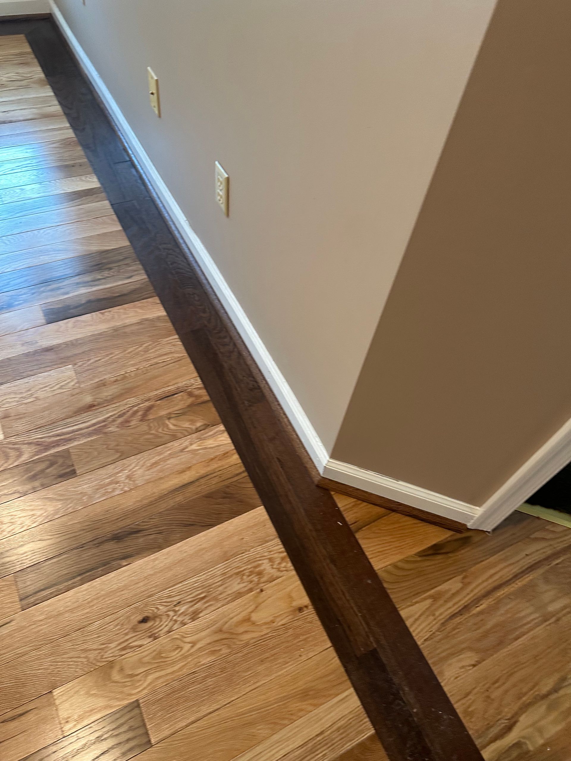 Hallway corner with wood floor, beige walls, white baseboard, and dark floor trim stripe