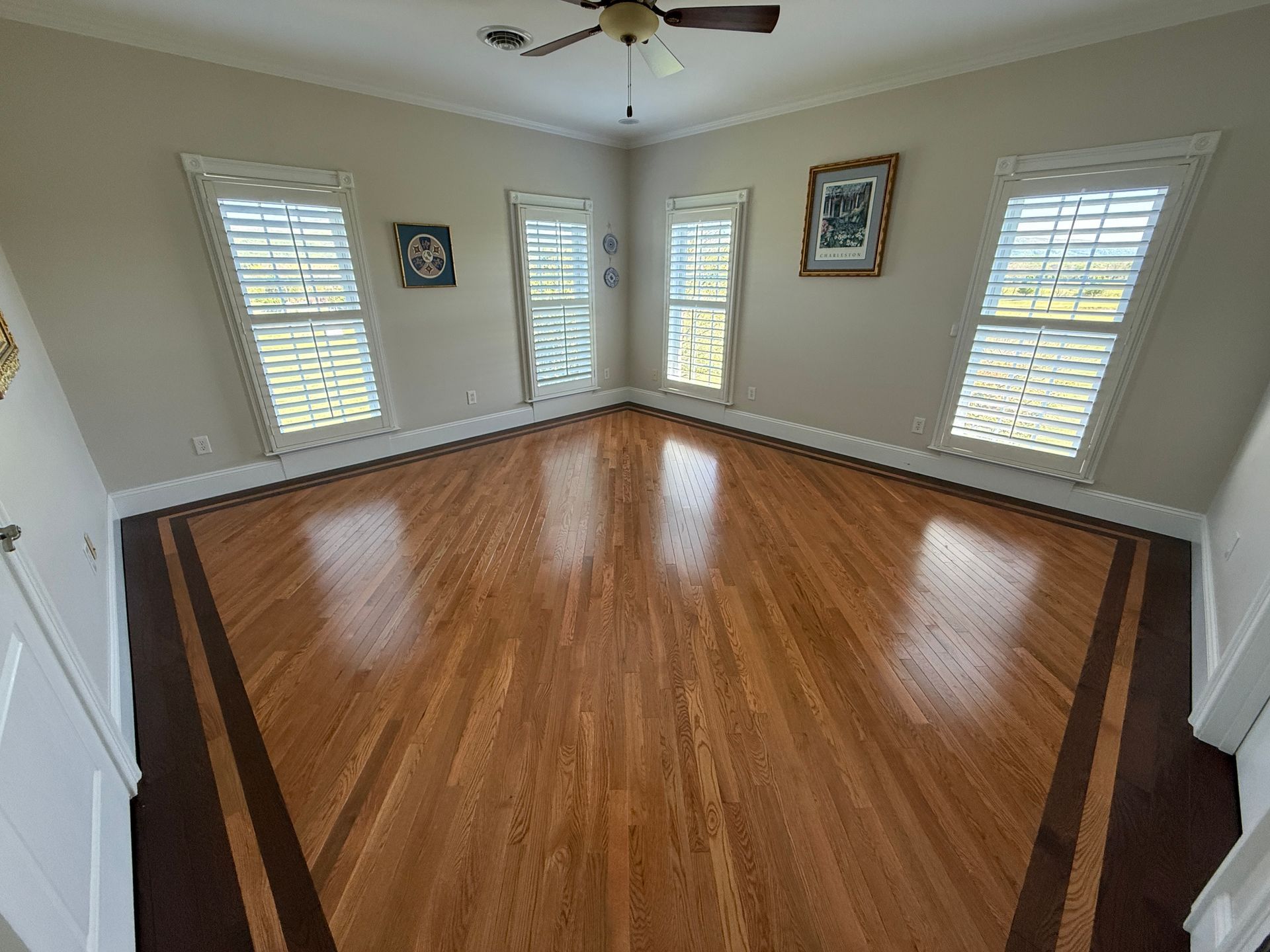 Empty living room with shiny hardwood floor, white walls, ceiling fan, windows, and framed wall art