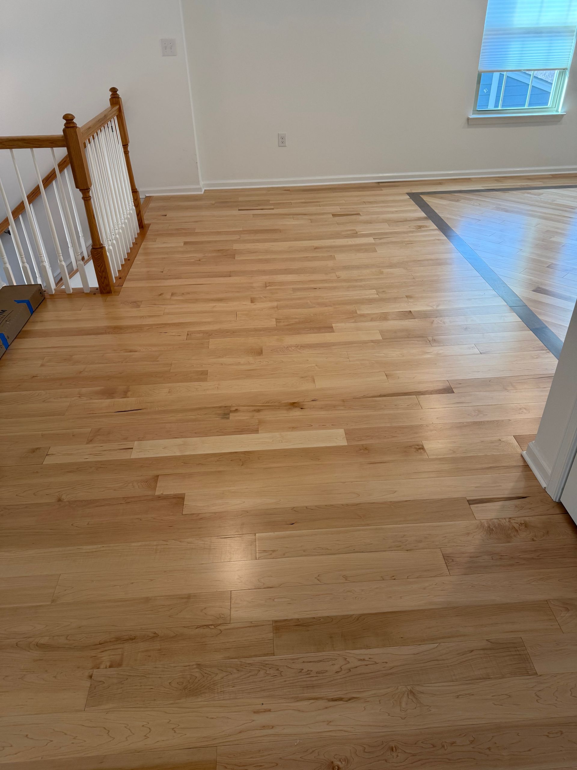 Empty light wood floor in a bright room with white walls and a window, next to a stair railing