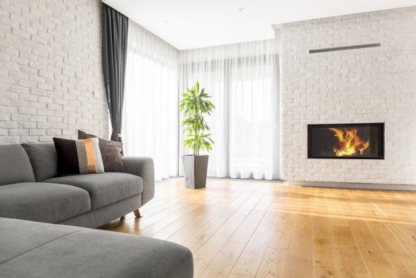 Bright modern living room with gray sofa, wood floor, potted plant, and lit fireplace under large windows