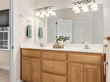 Bathroom vanity with light wood cabinets, white countertop, mirrors, and overhead lights
