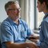 Man in blue shirt, doctor in scrubs converse in room, possibly medical exam.