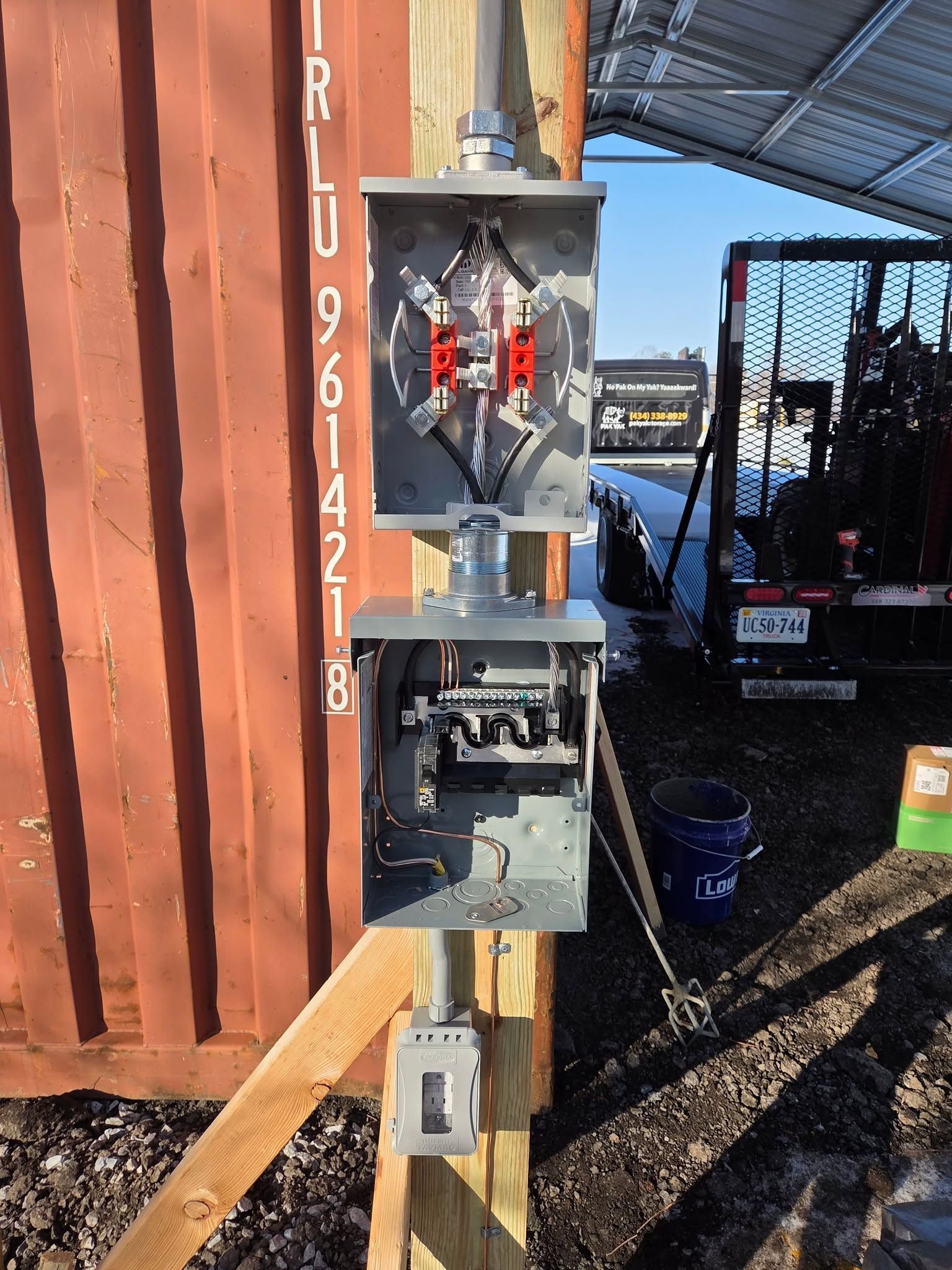 Electrical meter box mounted on a rusty shipping container, with exposed wiring and a forklift in the background