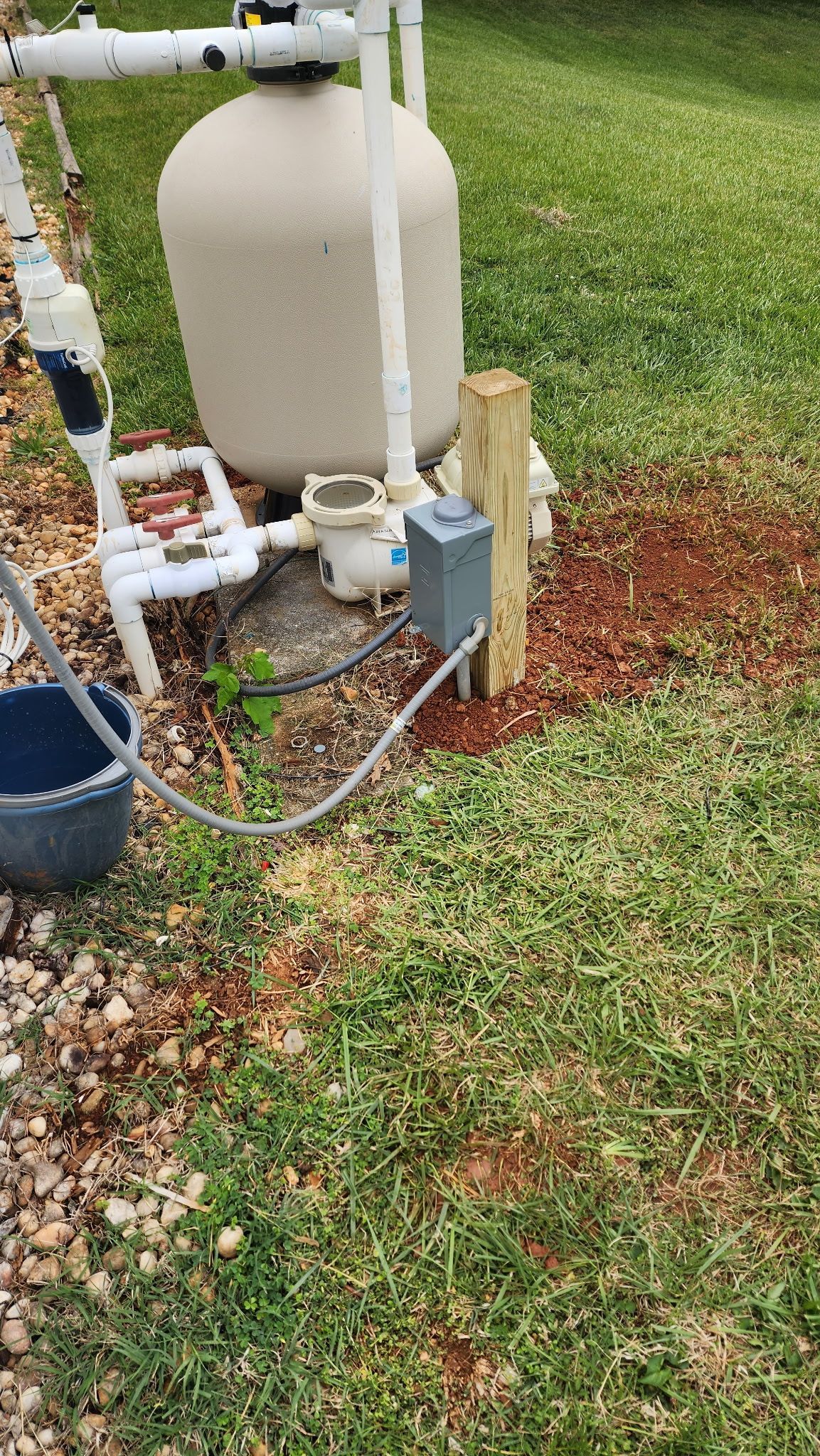 Outdoor pool equipment with white tank, pipes, and blue pump on gravel beside a grassy lawn
