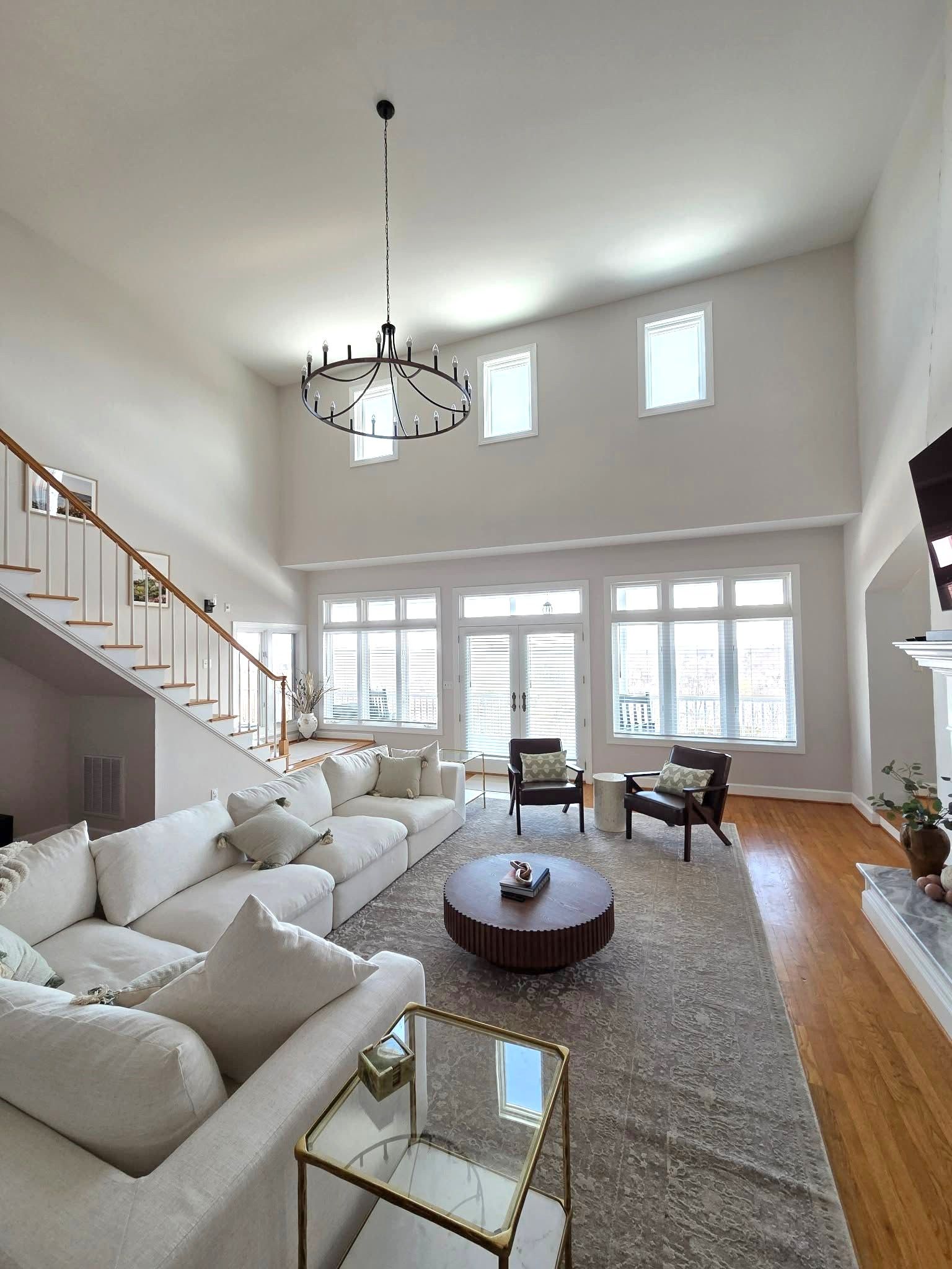 Bright, airy living room with white sofas, staircase, chandelier, and large windows.