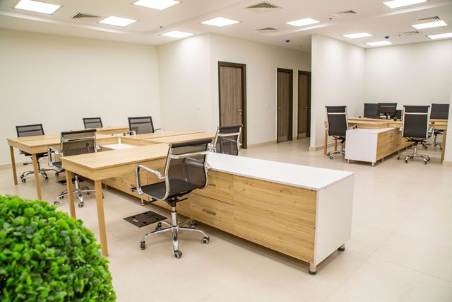 Bright open office with desks, chairs, computers, and a white central table under ceiling lights.