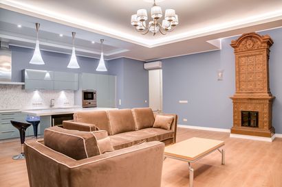 Modern open-plan living room with tan sectional sofa, chandelier, kitchen, and ornate wooden fireplace.