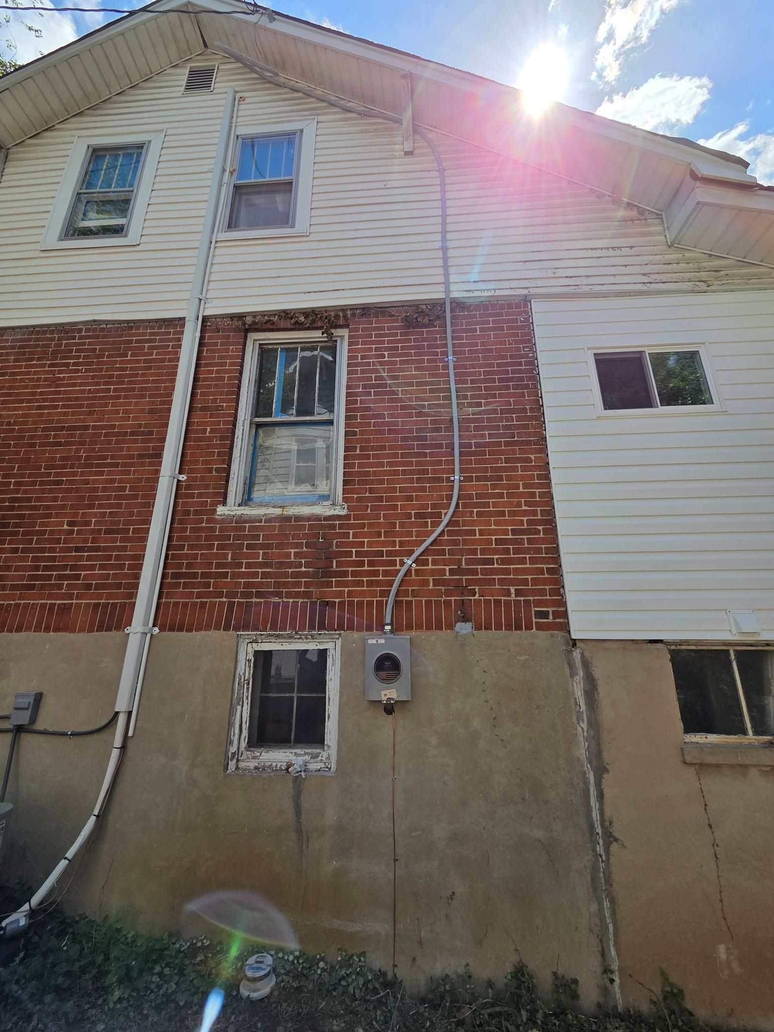 Back of a brick and white siding house with windows, utility meter, and bright sunlight flare