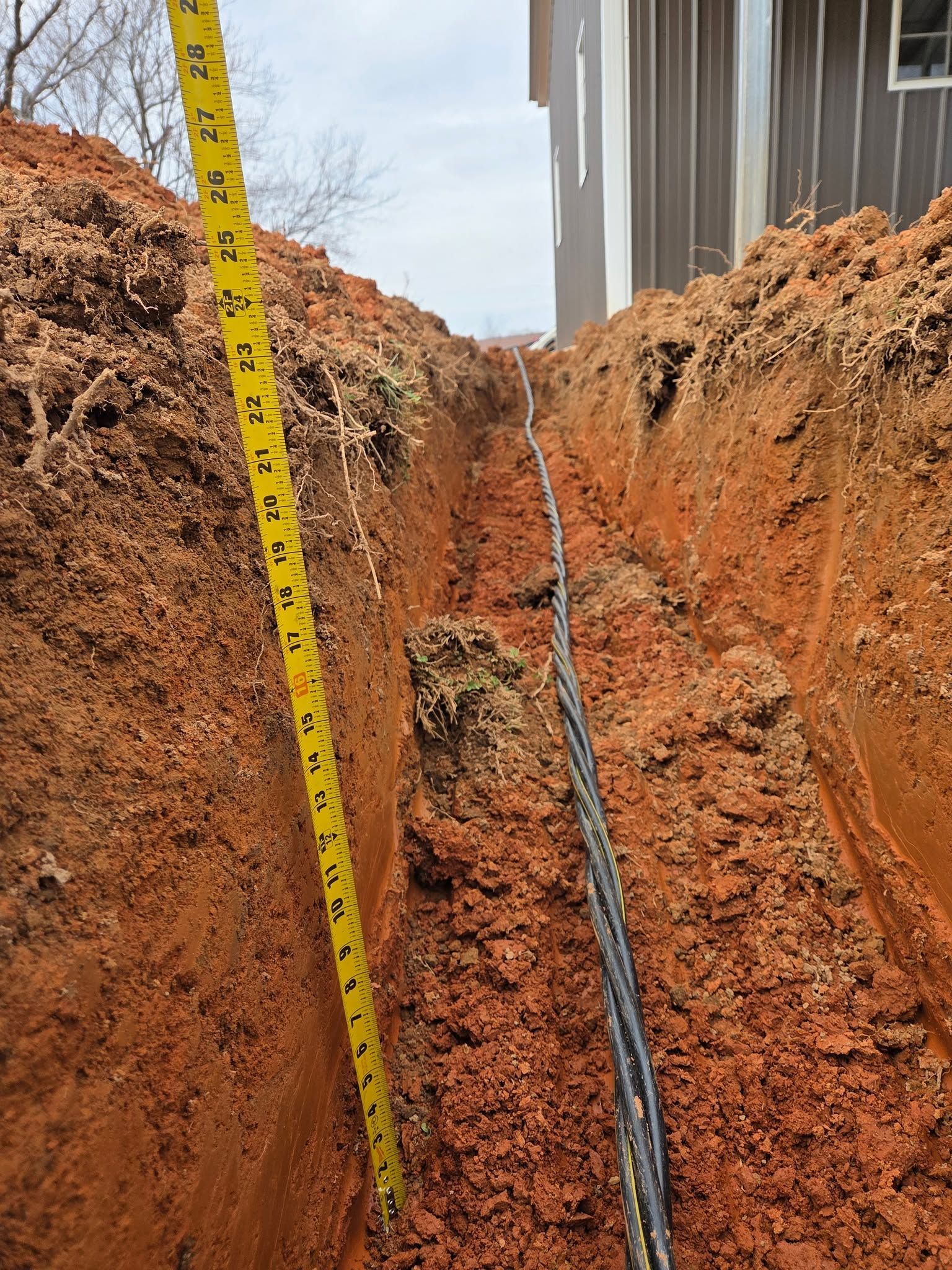 Trench with a yellow measuring tape beside exposed black cable and an orange dirt wall.
