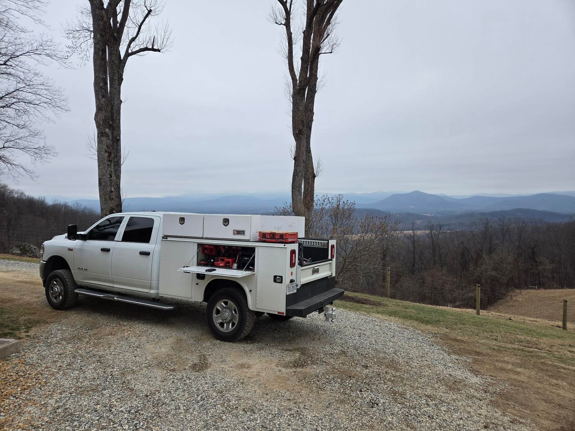 White utility truck parked on a gravel overlook between two trees, with mountains and a cloudy sky beyond.