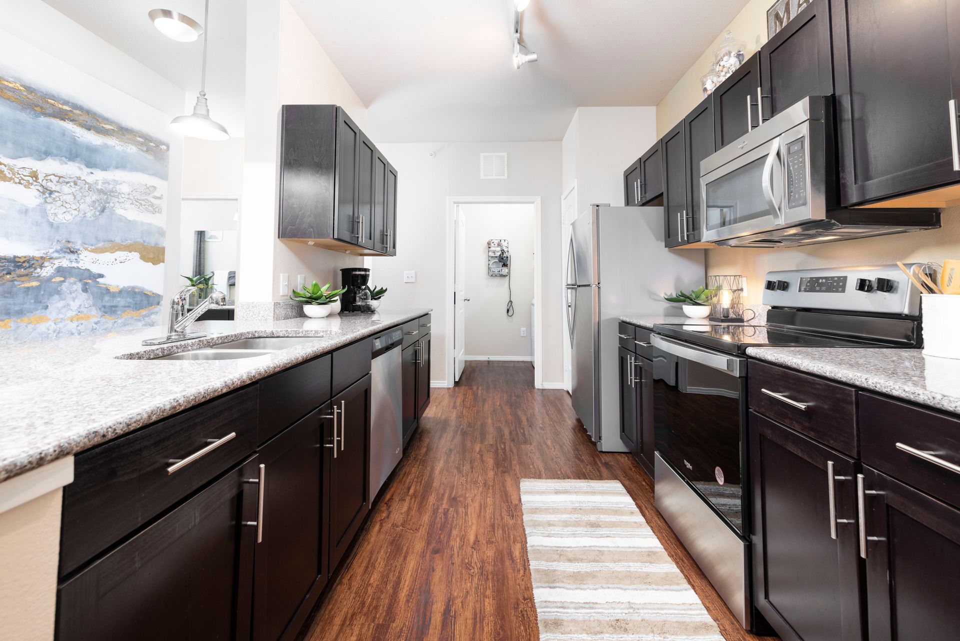 A galley-style kitchen with dark brown cabinets, granite countertops, stainless steel appliances, and wood flooring.