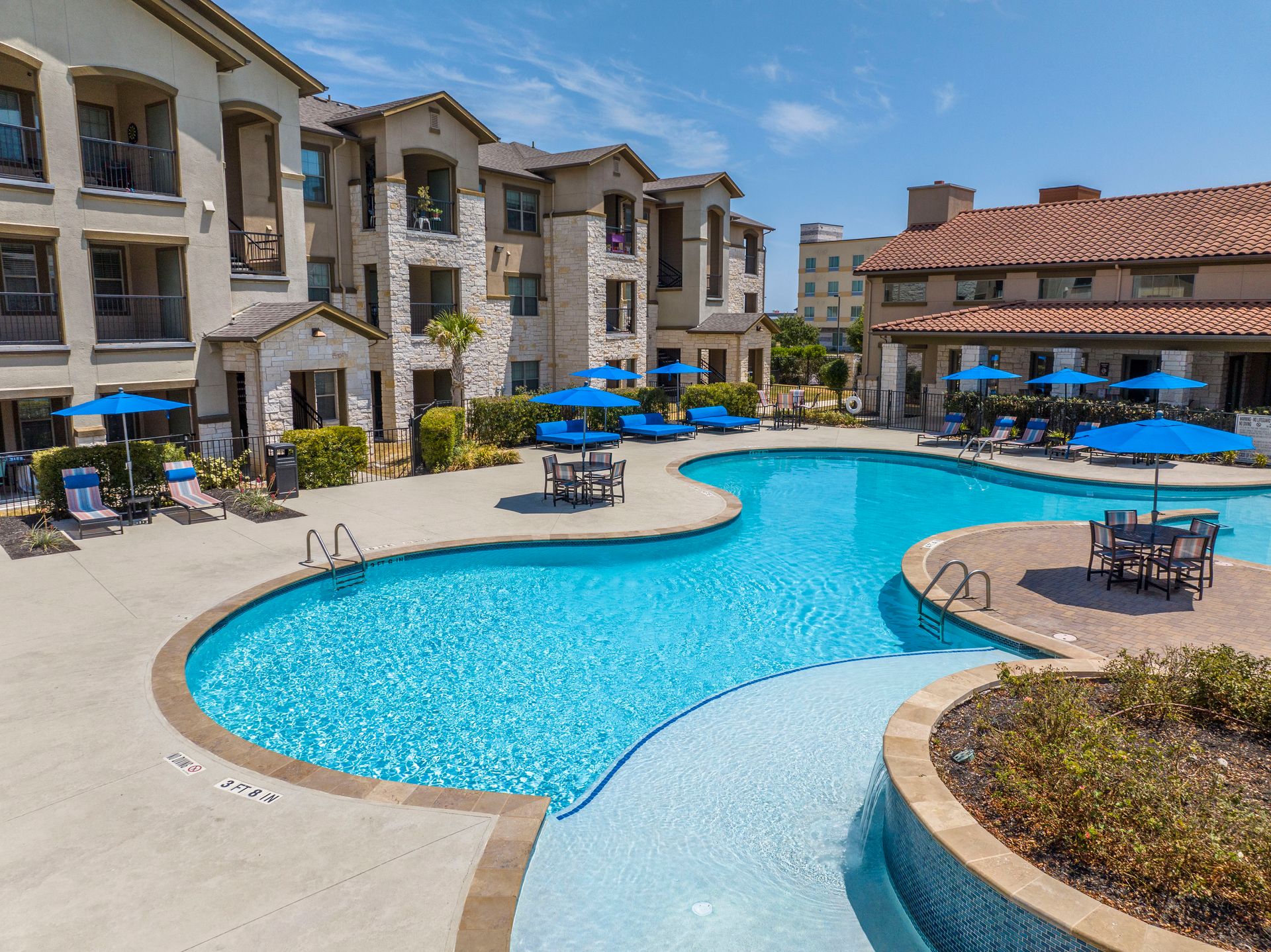 Sun-drenched courtyard featuring a curved swimming pool, blue umbrellas, lounge chairs, and an apartment building.