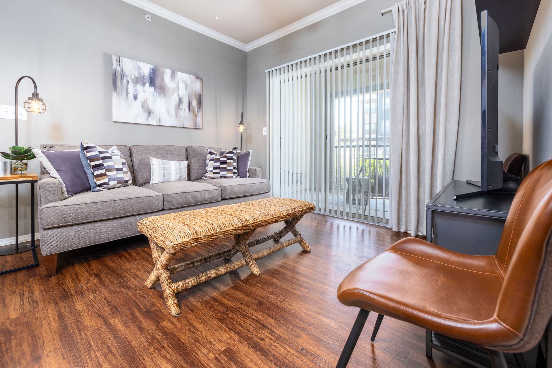 A living room with a gray sofa, a woven bench coffee table, and a brown leather chair on wood flooring near patio doors.