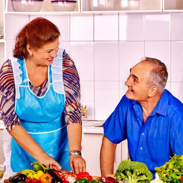 A woman in a blue apron and a man in a blue shirt smiling while preparing vegetables together in a kitchen.