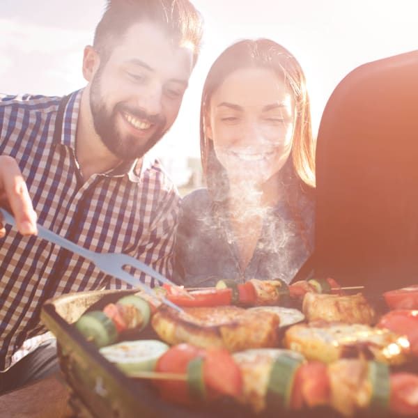 A smiling pair grills vegetable and meat skewers on an outdoor barbecue, smoke rising from the hot food.