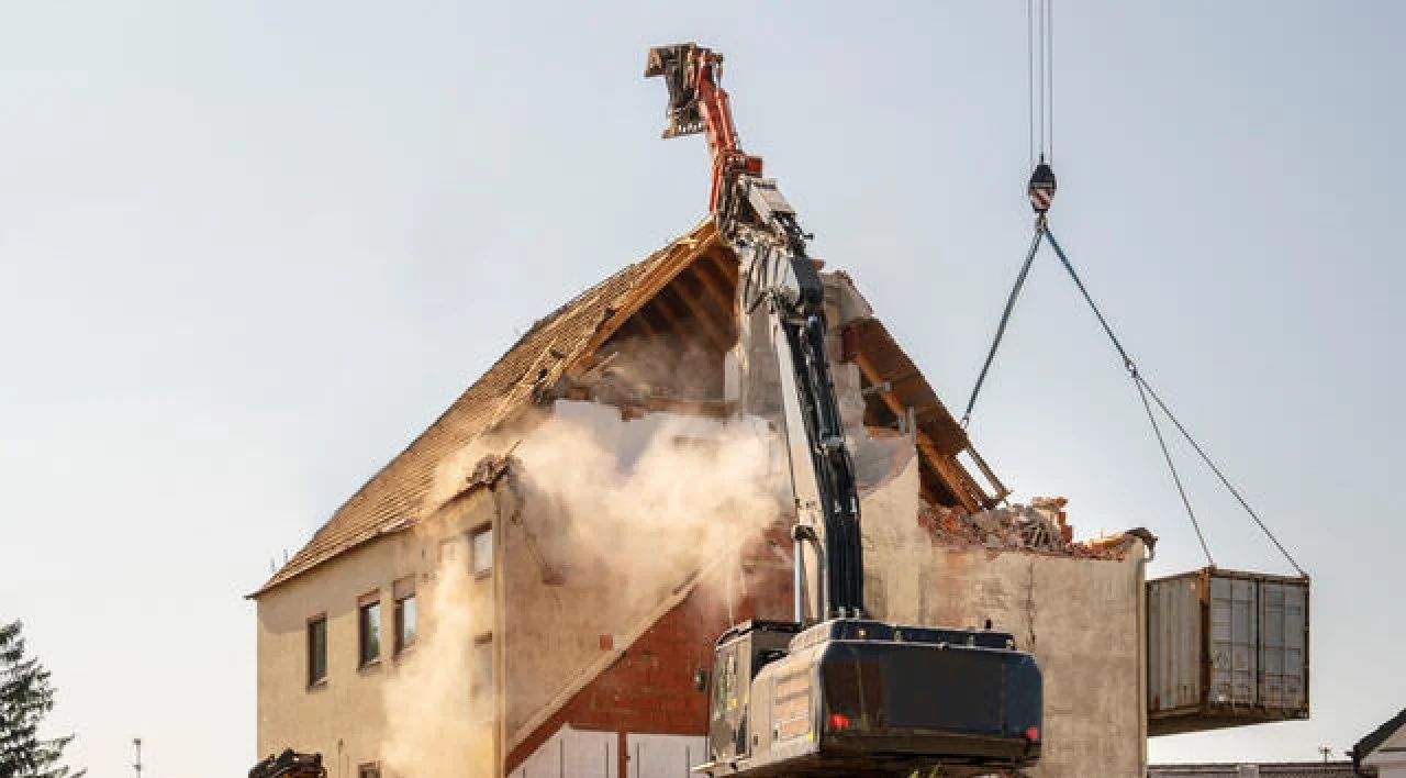 Demolition excavator tearing down a smoke-filled house, with debris hanging from a crane