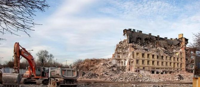 Demolition excavators clear rubble beside a damaged multi-story building under a cloudy sky.
