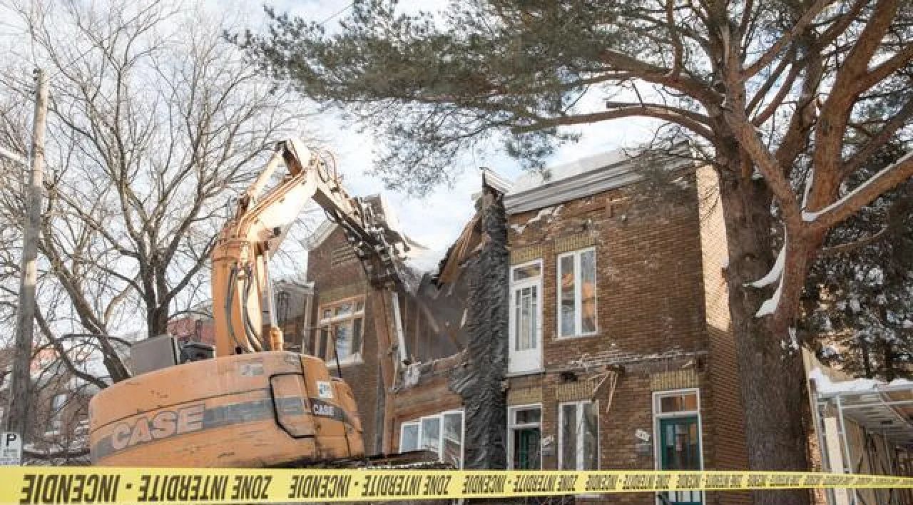 Excavator demolishes brick house behind yellow caution tape in winter.
