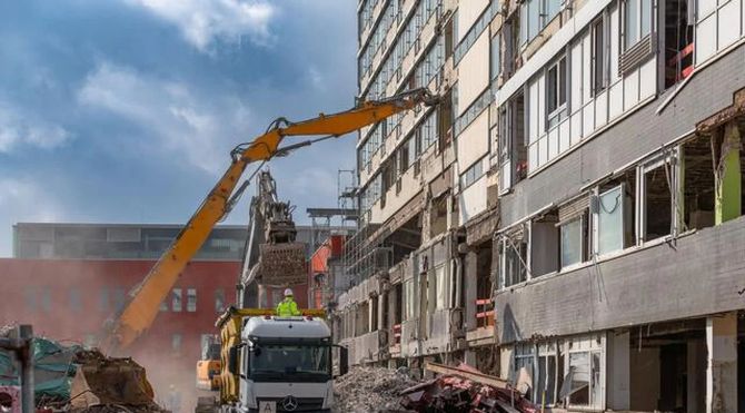 Excavator demolishes a building beside a truck in a rubble-filled construction site