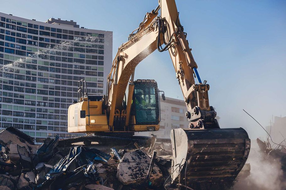 Excavator demolishing debris beside a tall glass building, with dust rising.
