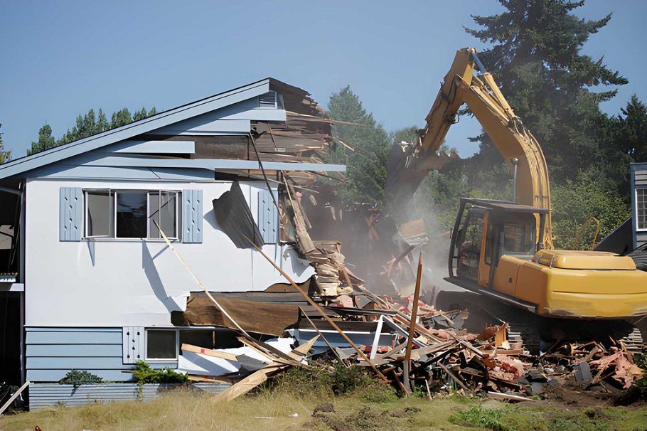 Excavator demolishing a white house, with debris falling beside the structure.