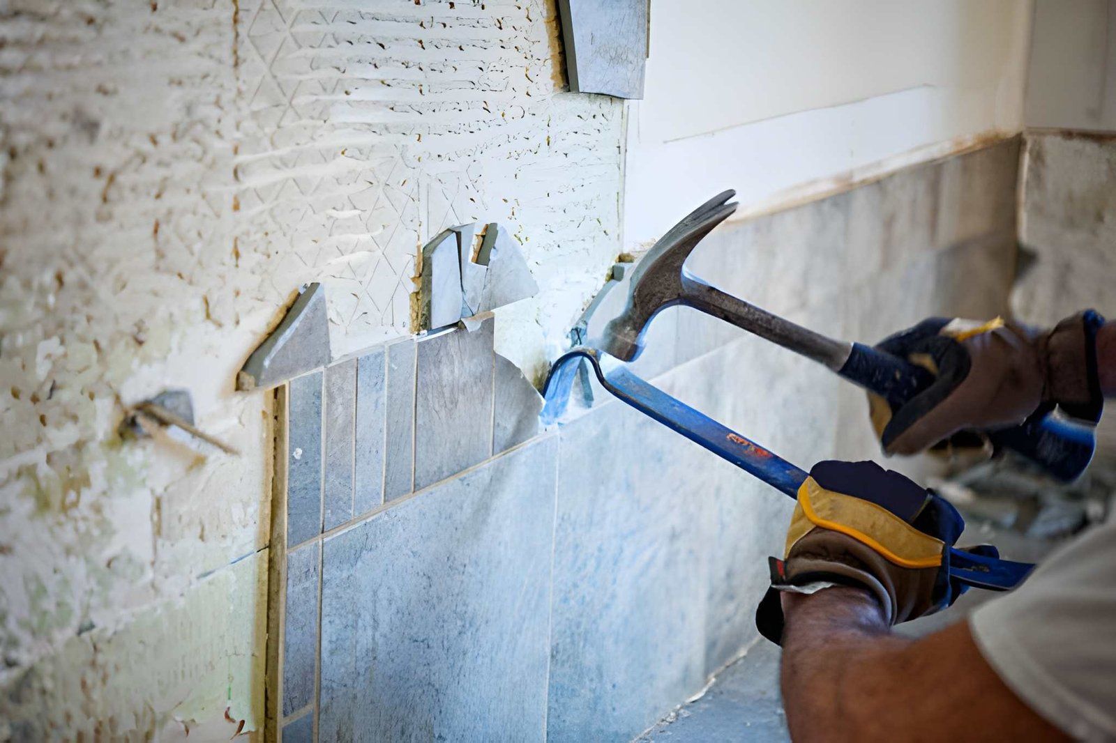 Worker using a hammer and pry bar to remove wall tiles during renovation