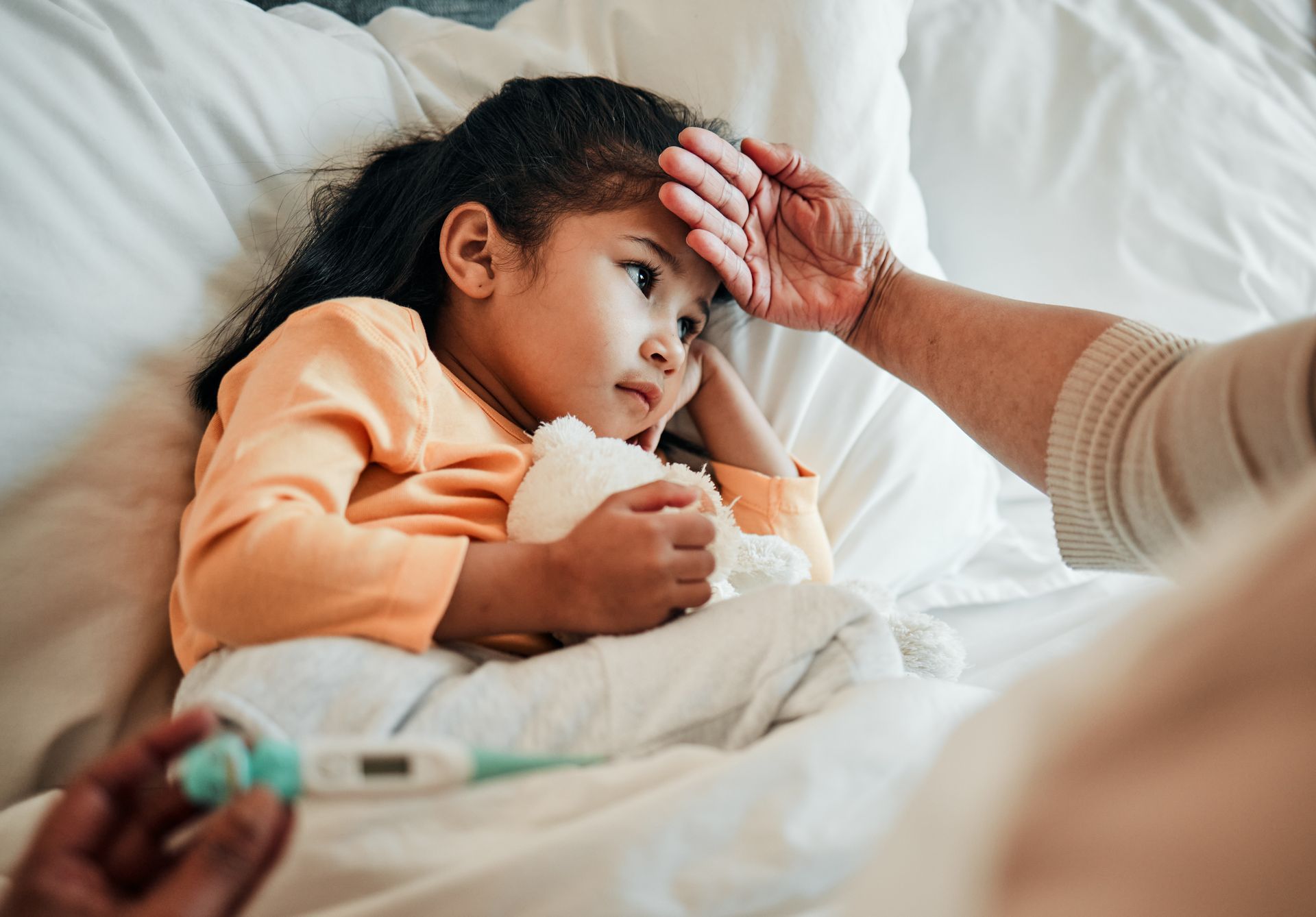 A young girl with a fever is in bed, while an adult checks her forehead with the back of their hand.
