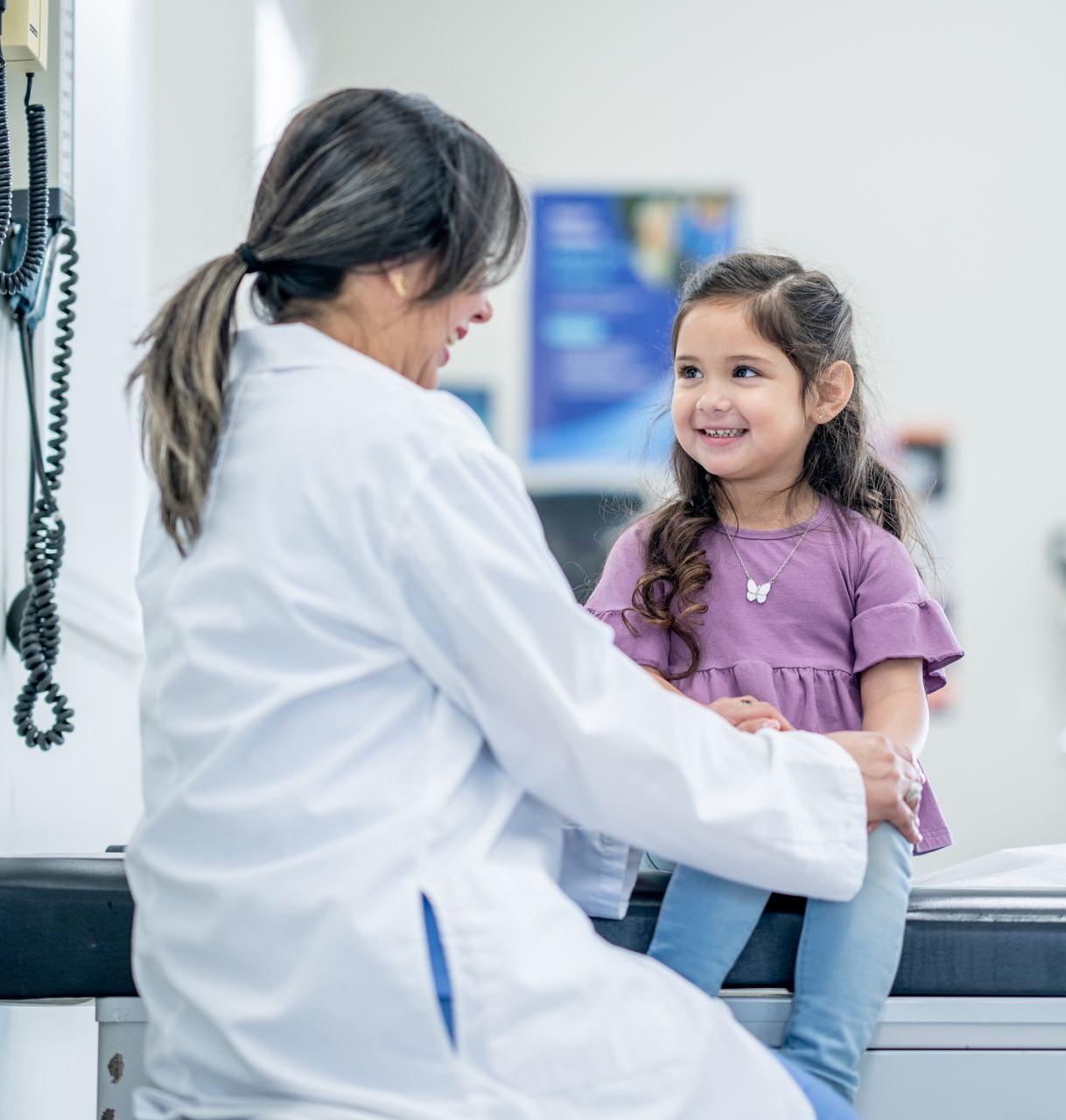 A female pediatrician is smiling at a young girl sitting on a medical table.