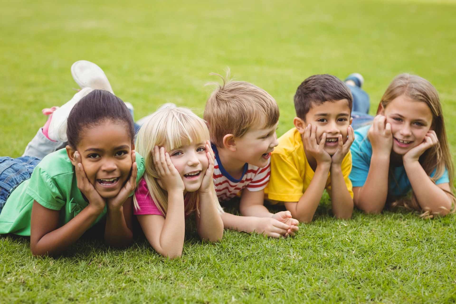 Several young children lying on their stomachs in a park with their hands on their faces. They are smiling.