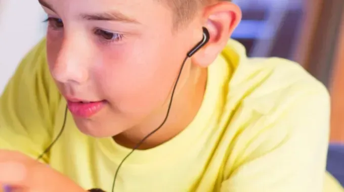 Boy with in-ear headphones wearing a yellow shirt looking down, indoors.
