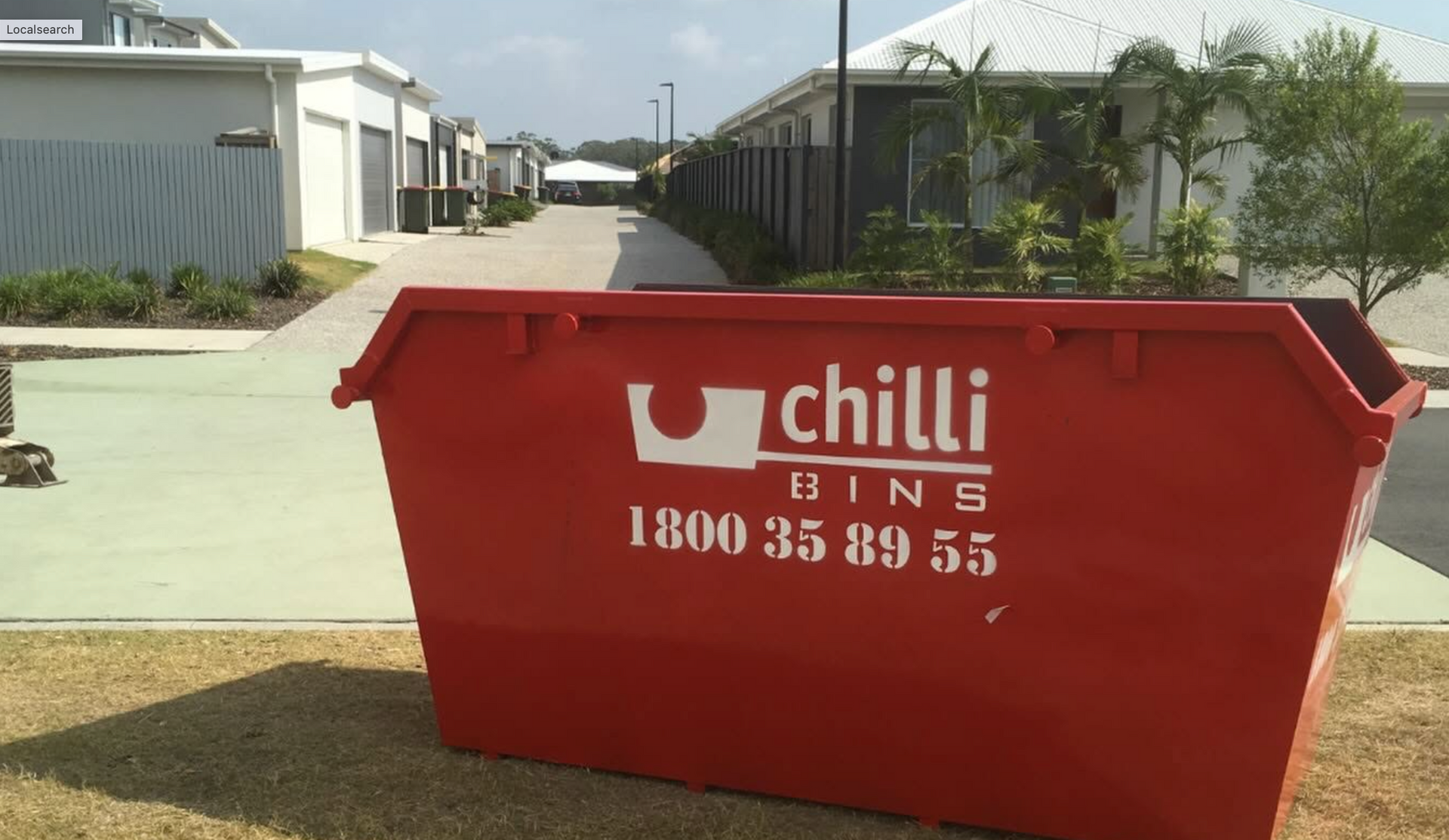 Red Chilli Bins skip bin on grass verge in front of modern townhouses along a residential street.