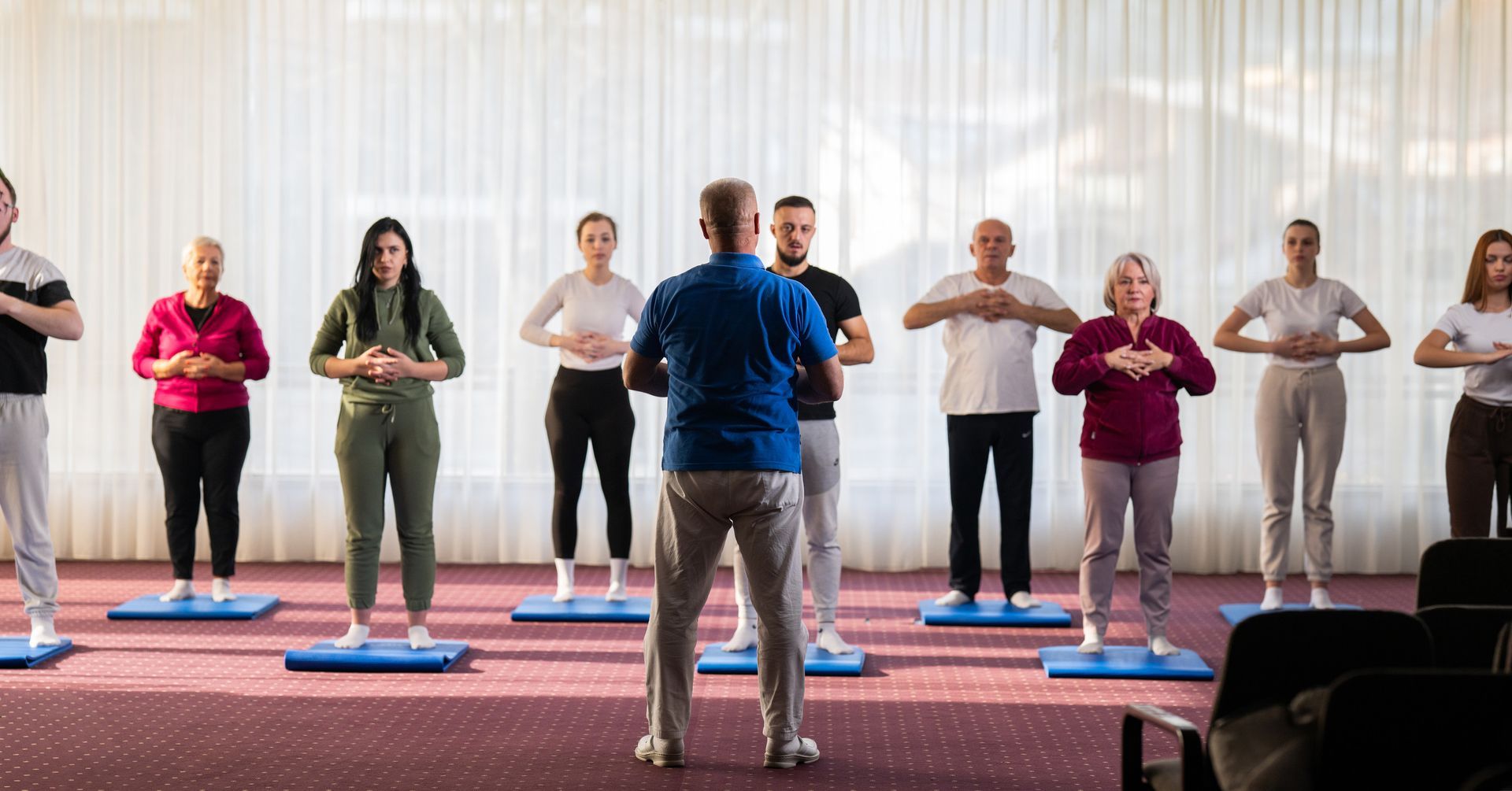 Instructor leading a group yoga class, all with hands in prayer pose, in a bright room.