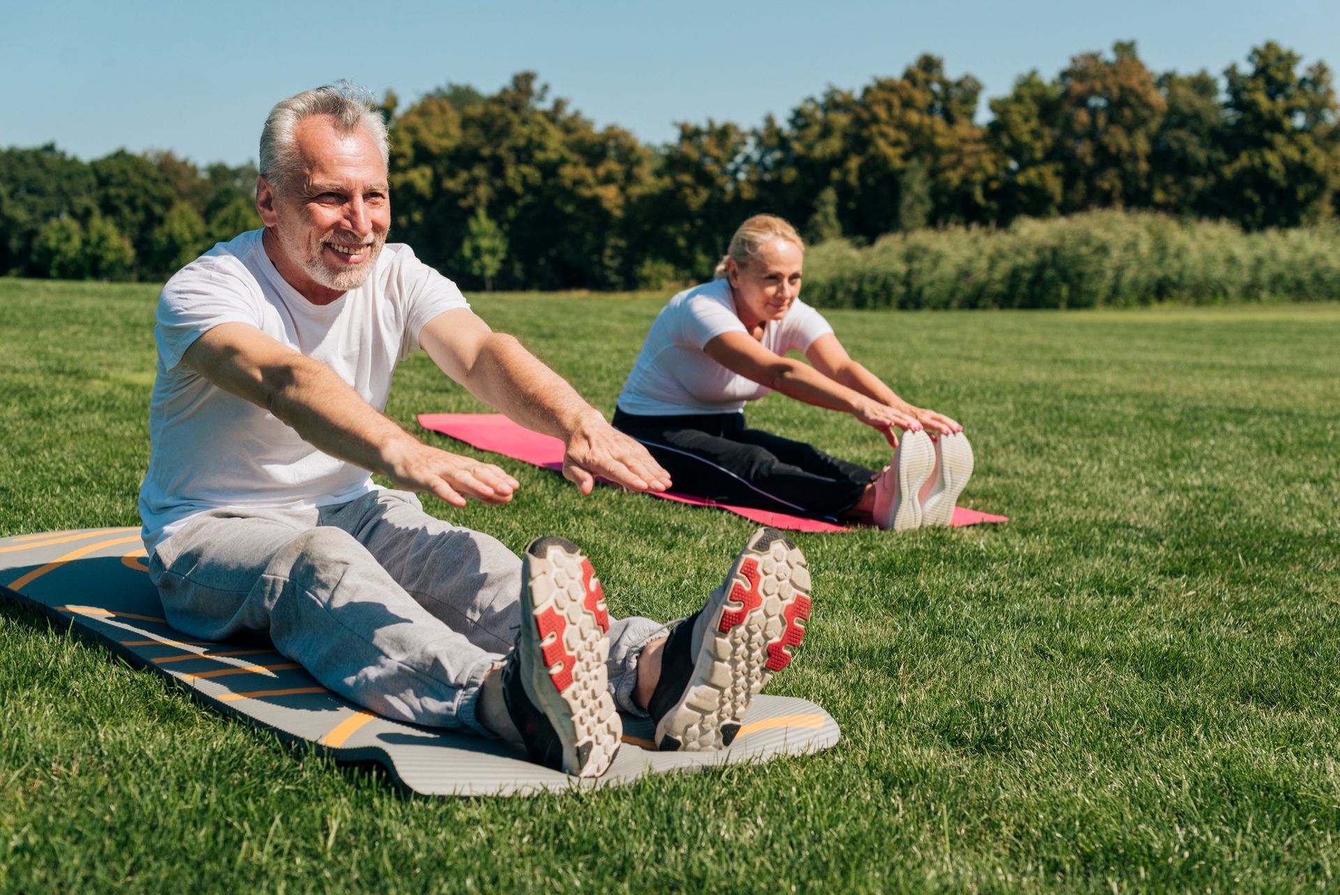 Two people stretching legs while sitting on mats in a grassy field. Sunny day.