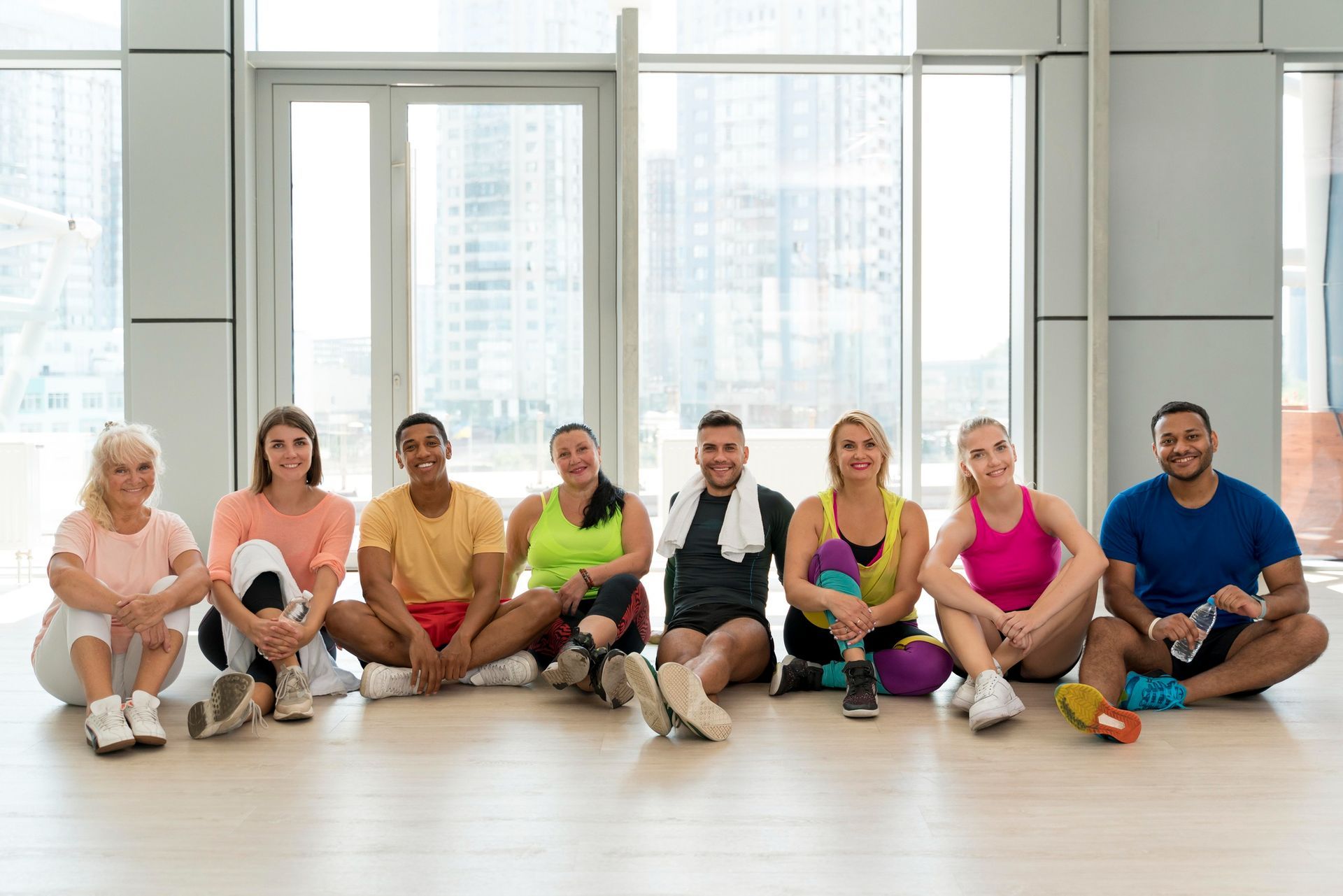 Group of people sitting on floor in a bright gym setting; smiling and resting after workout.