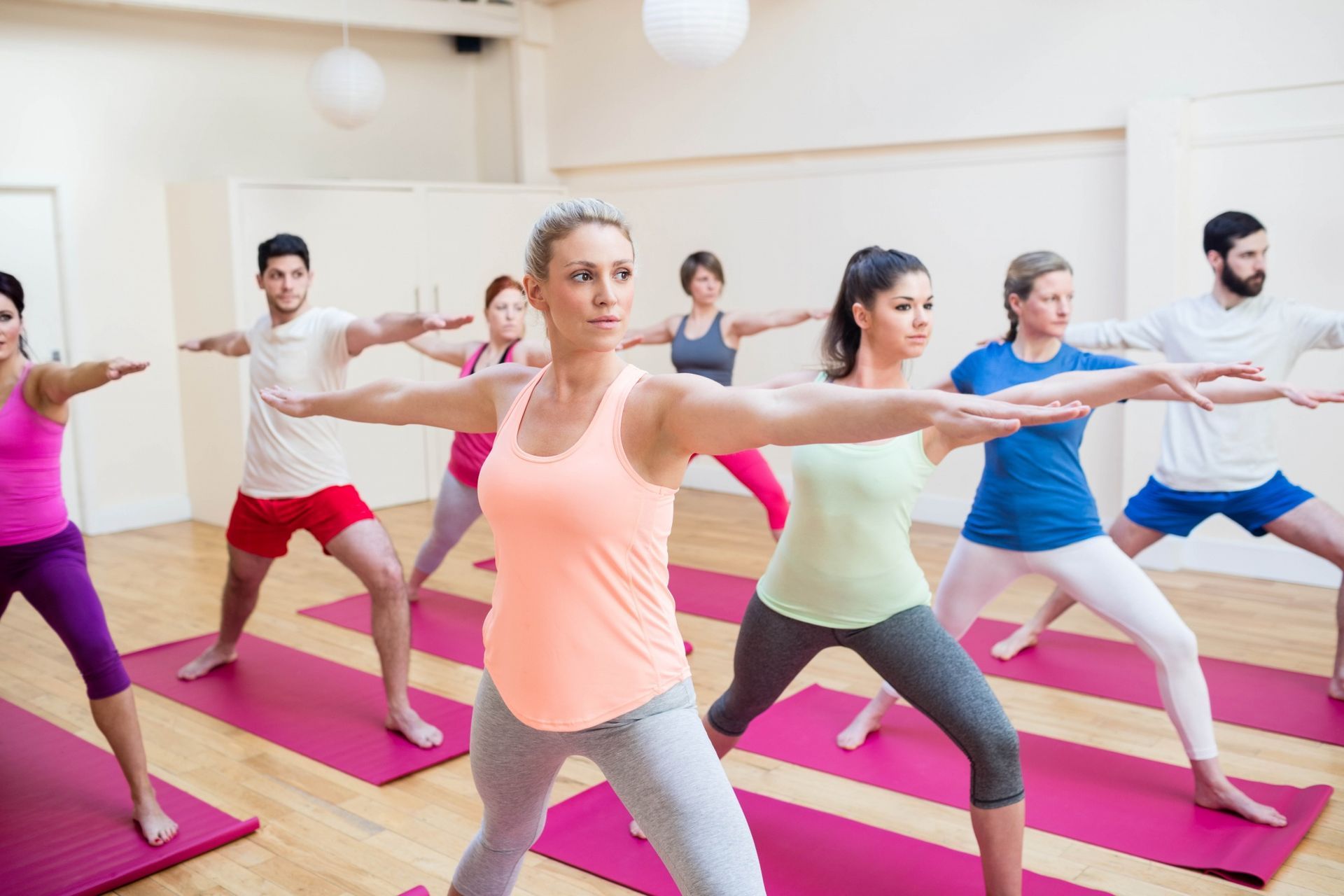 People in a yoga class, performing warrior pose on pink mats. Brightly lit studio.