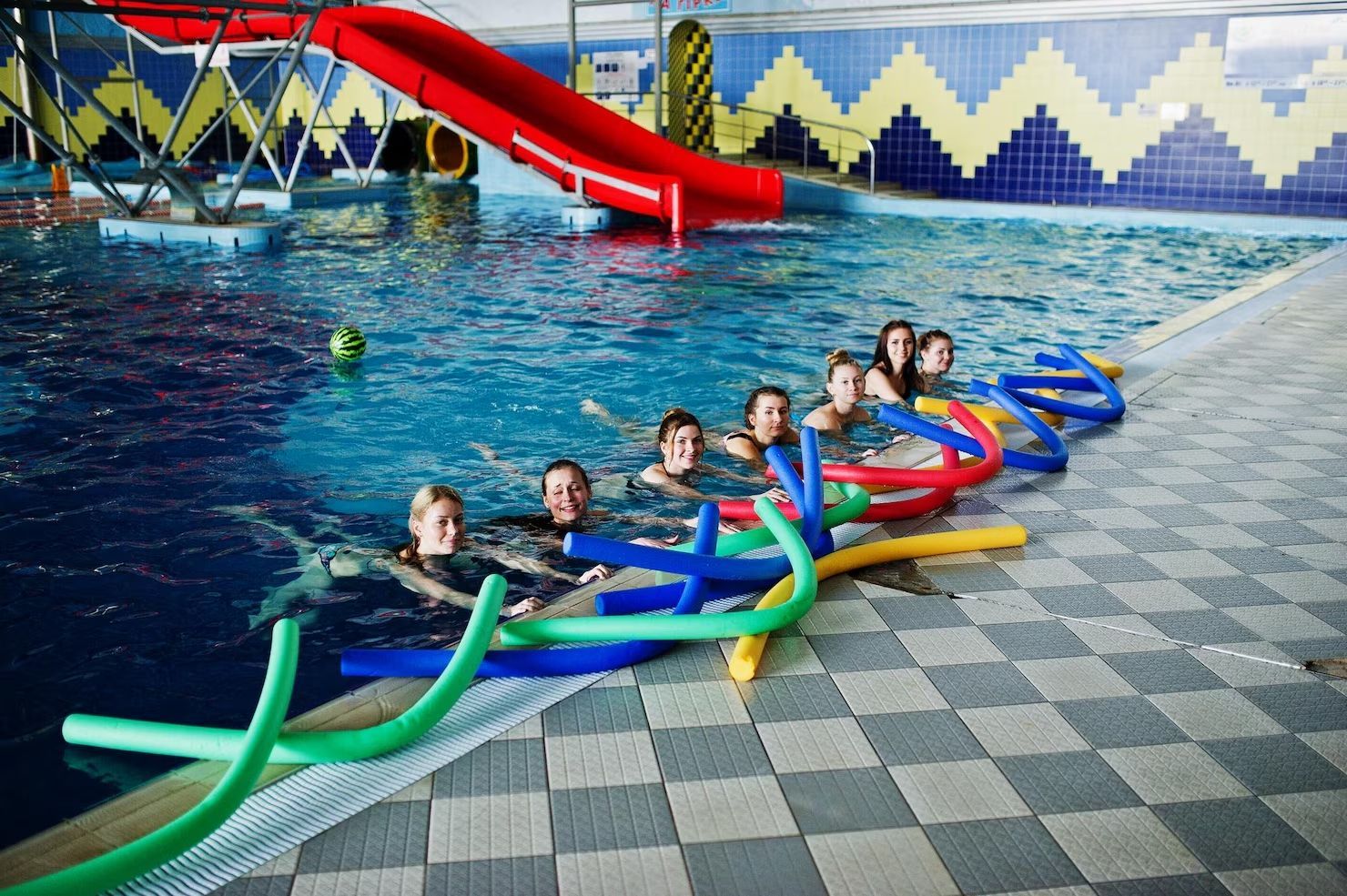 People in a pool with pool noodles, near a red slide in a blue-tiled indoor space.