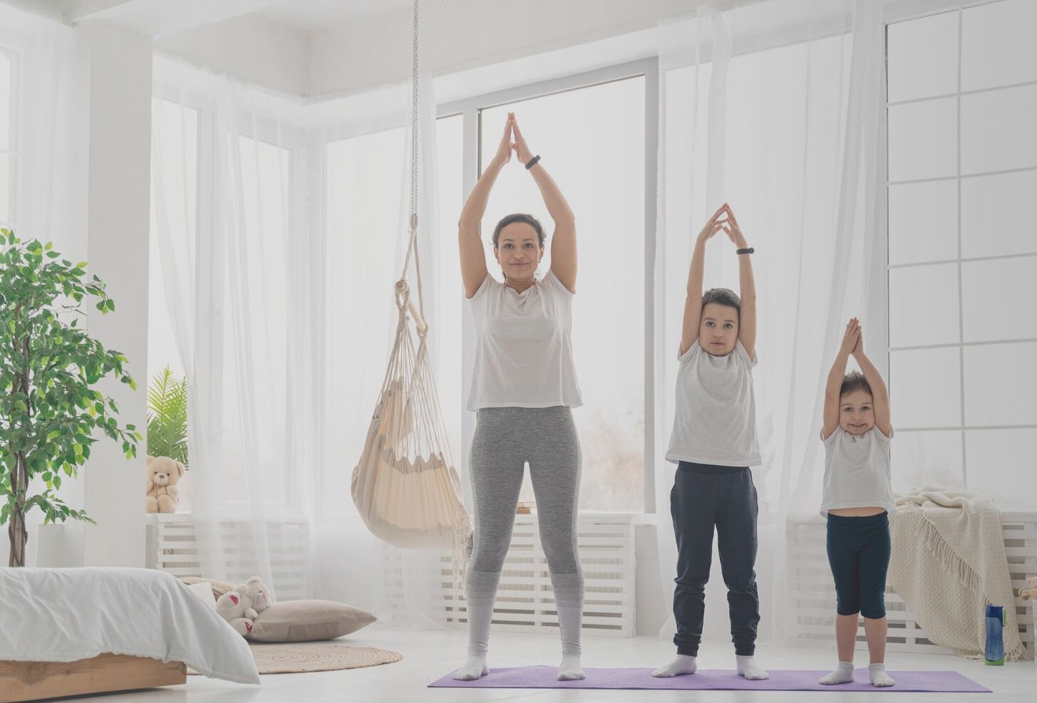 Woman and two children doing yoga with arms raised, in a bright room.