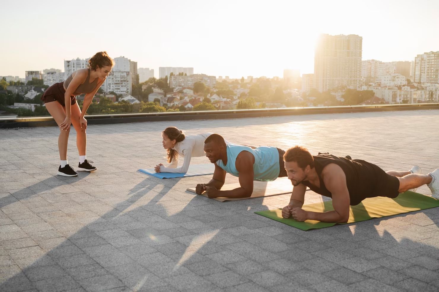 People doing planks on a rooftop under supervision of a person, cityscape in background.