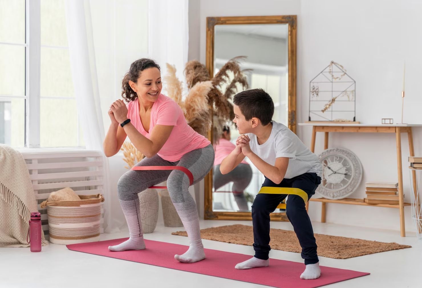 Woman and child squatting with resistance bands indoors on a pink mat.