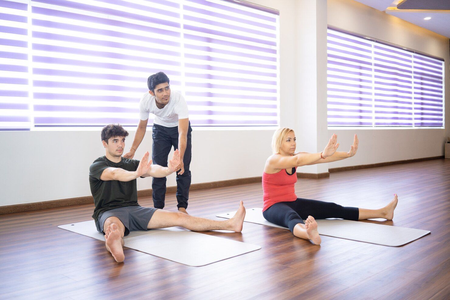 Three people doing floor exercises in a studio; instructor assisting two seated clients, all with arms extended.