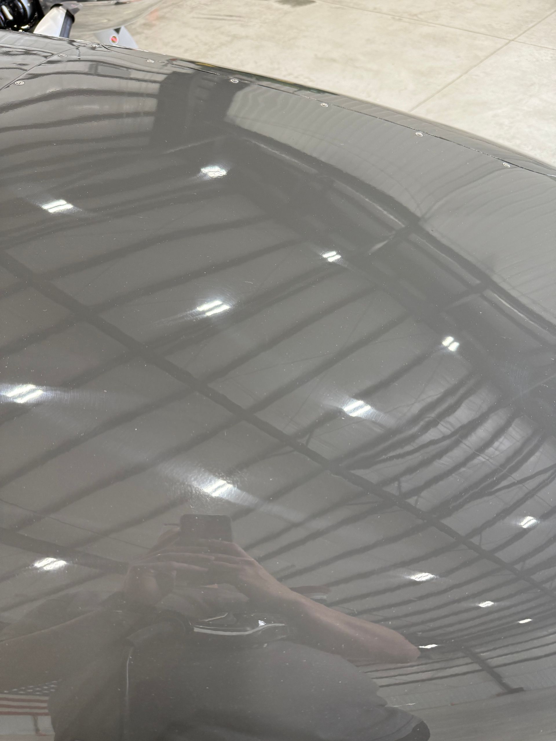 A dark gray airplane wing reflects a ceiling with lights. A person is reflected in the wing.