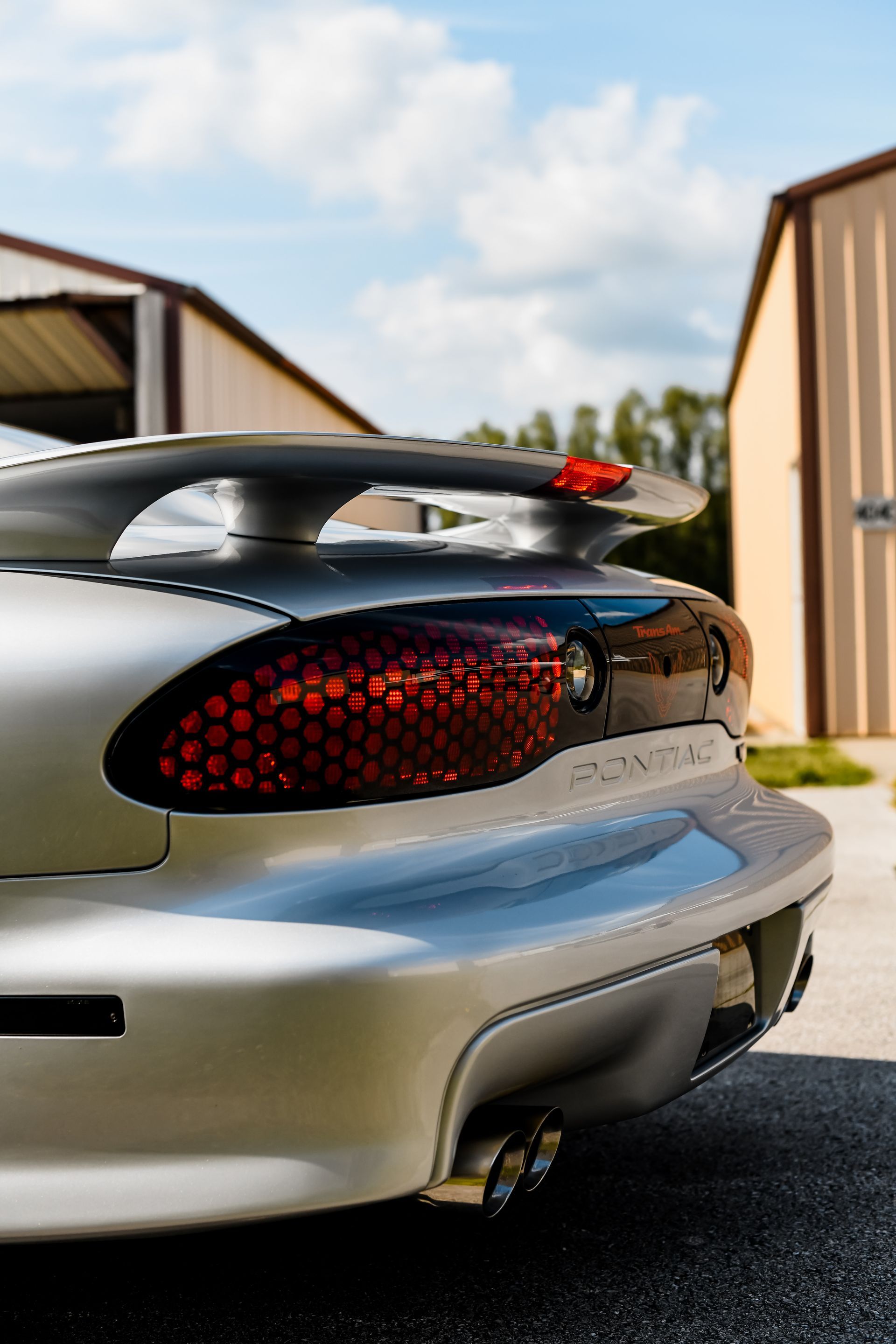 Silver Pontiac Firebird rear detail, with spoiler, taillight, and exhaust, in front of a building.