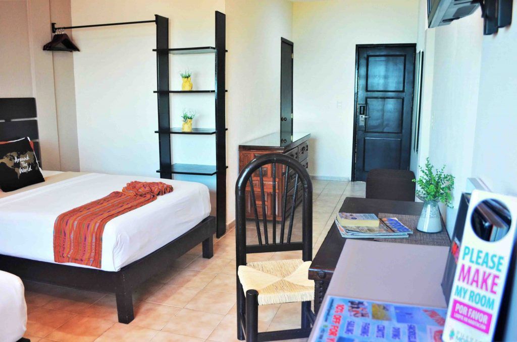 Hotel room with bed, chair, desk, shelving unit. Orange blanket, black wood furniture, white walls.