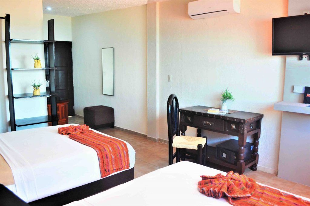 Hotel room with two beds, desk, TV, and decorative shelving. Bright, neutral tones.