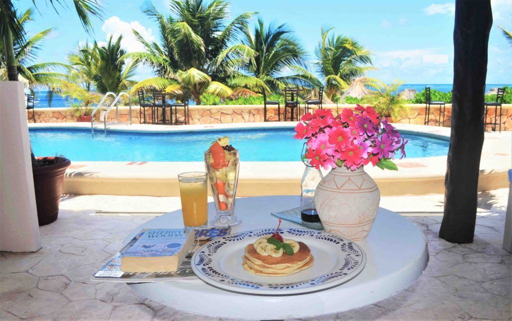 Breakfast by a pool: Pancakes, fruit, juice, and flowers on a table overlooking a pool and palm trees.