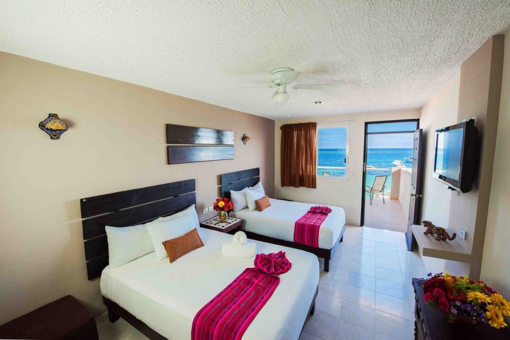 Hotel room with two beds, ocean view. Beige walls, white bedding, pink runner, TV, balcony.