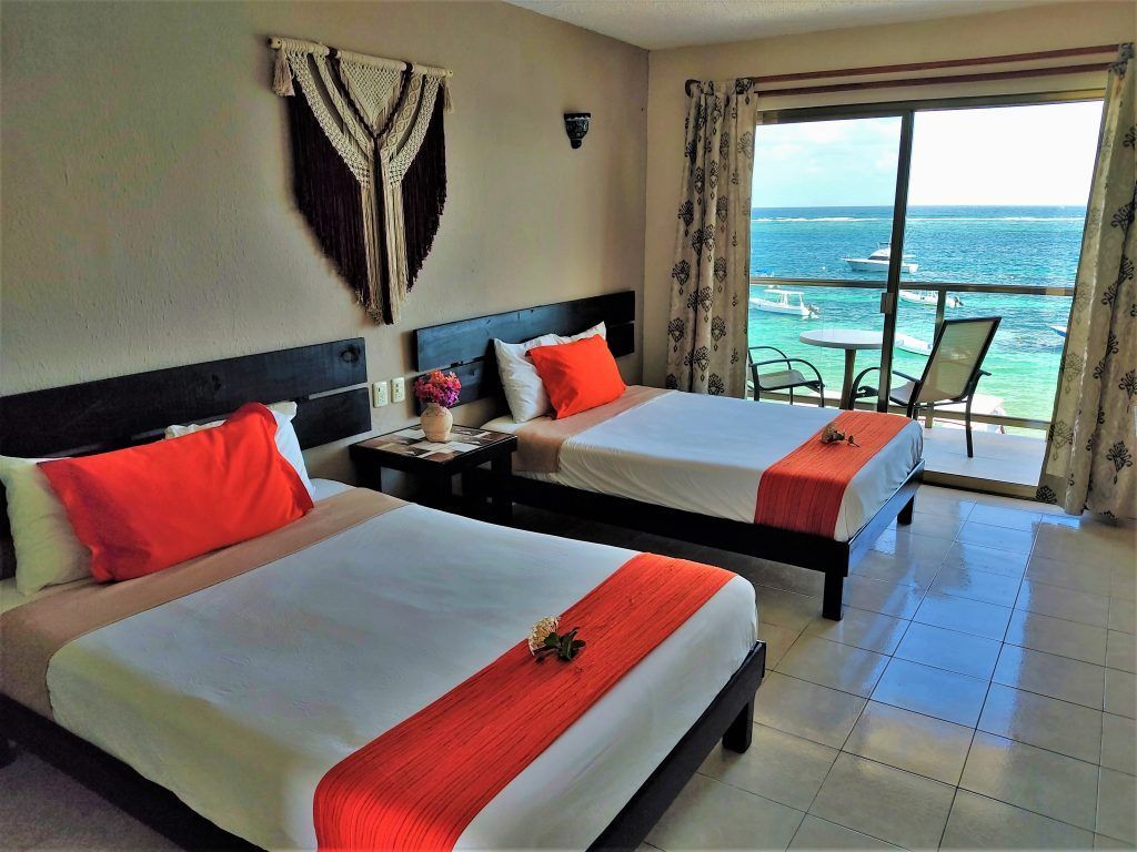 Two beds in a room, ocean view. Beds have orange pillows and runners, balcony with table and chairs.